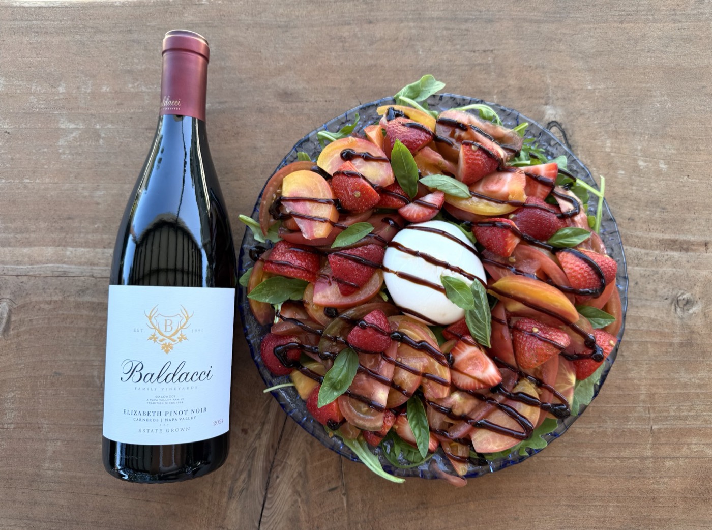 Overhead view of a strawberry burrata salad on a blue glass platter beside a bottle of Baldacci Elizabeth Pinot Noir